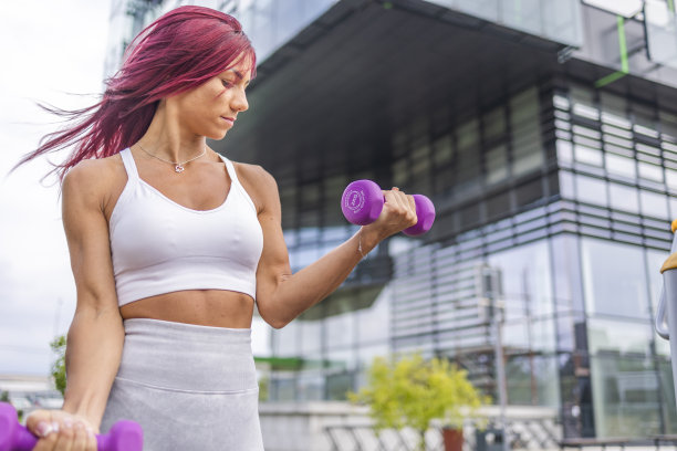 漂亮的粉色头发的健身女人在公司大楼附近的公园里举哑铃。图片下载