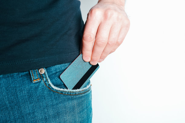一个年轻人从他的蓝色牛仔裤口袋里掏出一张信用卡。一个男人的手从裤子里掏出一张支付卡的特写，背景是浅色的。图片下载