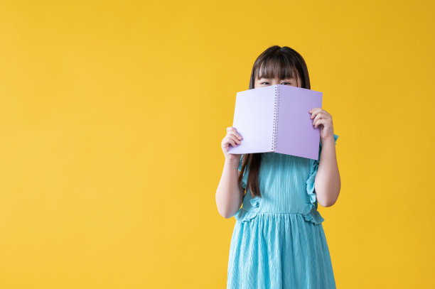 一个可爱的年轻亚洲女孩正在发现或隐藏她的脸后面的一本书在她的手中。图片下载