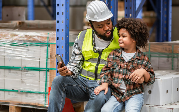 非洲的父亲或父亲在仓库或工厂工作时，用爱拥抱他的小儿子，带着幸福的微笑，戴着安全安全帽，一起教学，一起交谈。教育，产业理念。图片下载