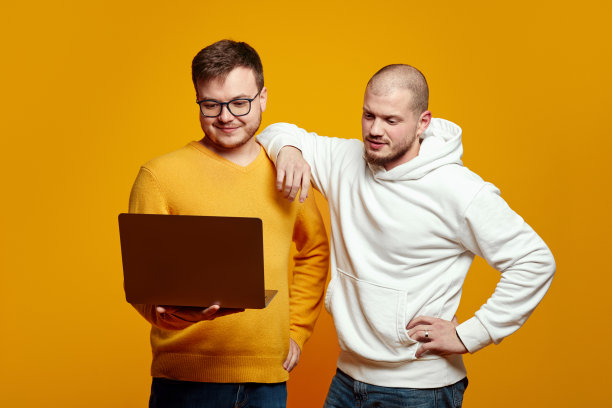 两个英俊的年轻人拿着笔记本电脑，看着笔记本，隔着黄色的背景图片下载