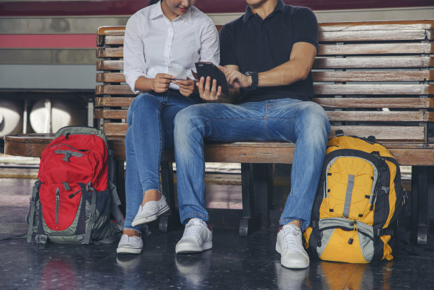 廉价旅行和生活方式的概念。年轻的旅行者男女(一对情侣)穿着运动鞋坐在火车站。亚洲背包客在等火车，用智能手机计划旅行。图片下载