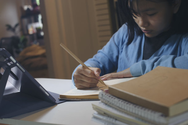 亚洲孩子在晚上用平板电脑做作业和学习作业。小女孩在家学习、读书、上网工作。亚洲小孩在日记本上写信。学生开心而微笑。教育的概念。图片下载