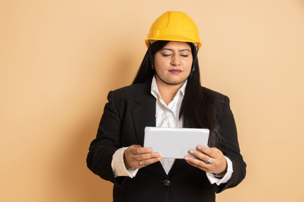 印度女工程师或建筑师，穿着黑色正装，戴着黄色安全帽或头盔，手持电子平板电脑，孤立在米色背景上。建筑的概念。副本的空间。图片下载
