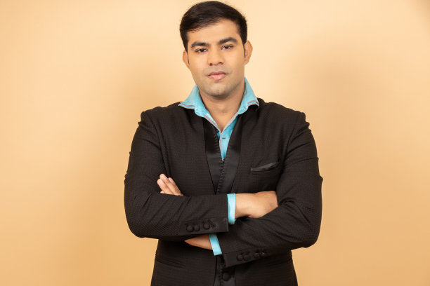 年轻英俊的印度男子穿着黑色正装，双臂交叉站在米色背景上。商业和企业概念。图片下载
