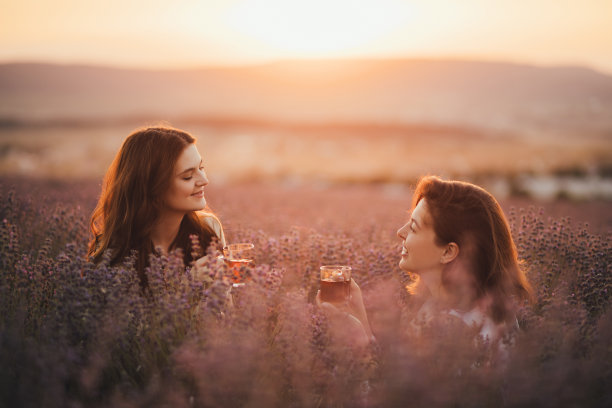 两个快乐的女人喝着健康的花草茶，坐在夕阳下美丽的薰衣草地里。图片下载