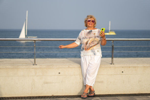 在一个阳光明媚的日子里，在海滩附近，一位白发苍苍的迷人老妇人微笑着读着智能手机上的信息。这位老妇人拿着手机享受着她的空闲时间和假期图片下载