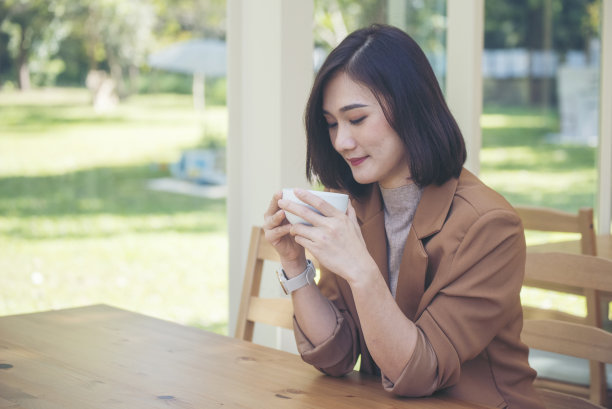 亚洲妇女在绿花园咖啡馆用手捧杯喝黑咖啡。年轻女子笑着脸爱喝咖啡。美女拿着新鲜的杯子在咖啡店喝黑咖啡。咖啡爱好者图片下载