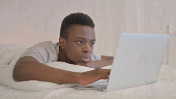 年轻的非洲男子趴在床上用笔记本电脑工作图片下载