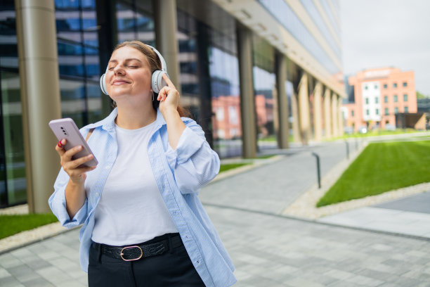 微笑的白人年轻女商人，戴着耳机听着播客电子书、音乐、歌曲、歌手、摇滚乐队，用手机选择音轨，走在城市街道上。高质量照片图片下载