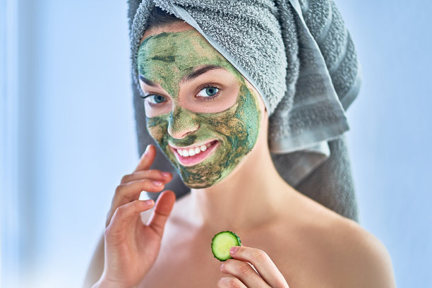 肖像微笑的快乐年轻健康的妇女与绿色清洁的脸粘土面膜和新鲜的黄瓜片在家里的水疗日。面部皮肤护理图片下载
