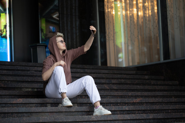 一名穿着连帽衫和白裤子的成年女子坐在外面的楼梯上，用她的手机自拍。图片下载