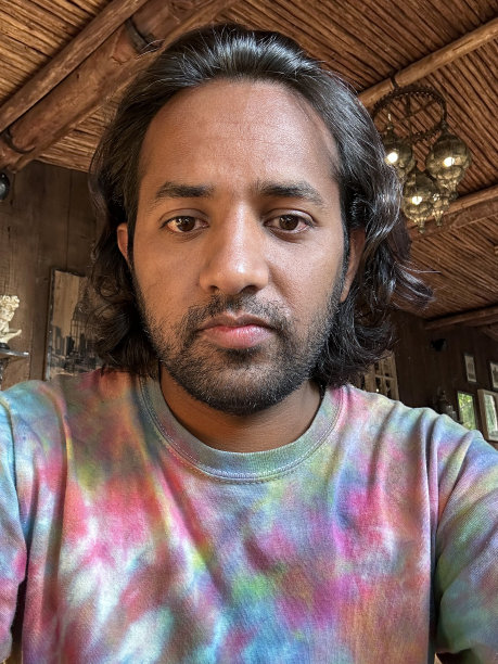 特写的印度男子深色胡子穿着休闲服装，自制，扎染t恤，懒洋洋地在餐厅酒吧，看着相机，棕榈叶屋顶和悬挂灯罩的背景，大头照，重点在前景图片下载