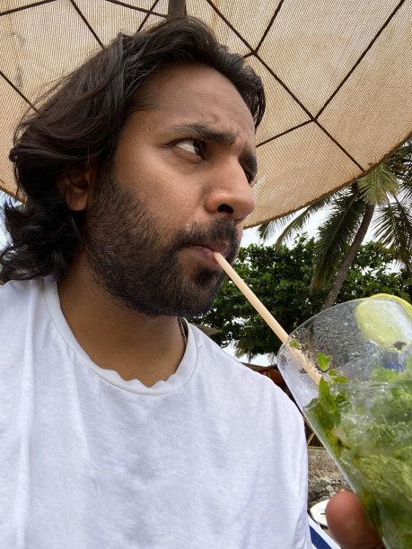 特写照片:一个印度人喝着一杯加冰、薄荷叶和柠檬片的莫吉托鸡尾酒，通过吸管，坐在沙滩餐厅的酒吧里，看着镜头，坐在遮阳伞下，聚焦于前景图片下载