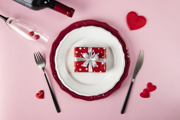 情人节浪漫的餐桌布置。礼物放在盘子里，粉红色背景的酒瓶和玻璃杯。餐厅约会图片下载