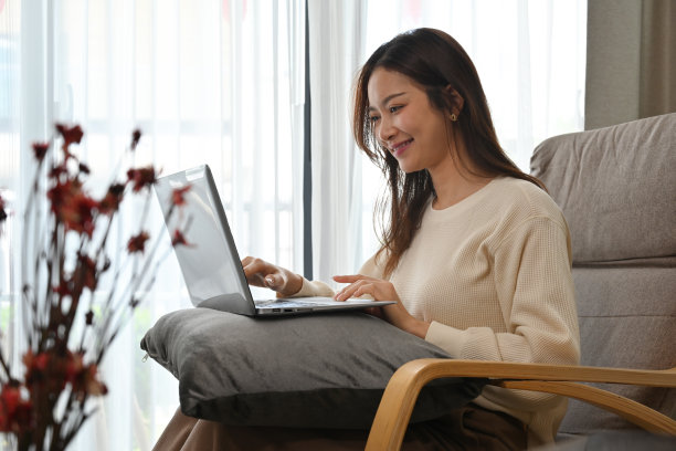 侧视图与年轻的亚洲妇女坐在扶手椅和使用笔记本电脑在家里。图片下载