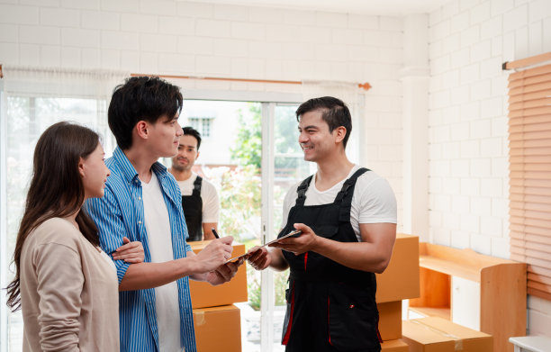 一对亚洲夫妇一边检查，一边把箱子和家具从一辆小货车上卸到一所有服务货物的新房子里，两名穿着制服的搬运工搬运箱子。搬家和送货的概念。图片下载
