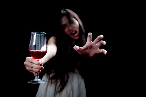 美丽的黑色长发女子手持红酒杯，黑色背景中的幽灵幽灵服装，恐怖的死人女性用恐怖的手拿着酒杯庆祝万圣节派对。图片下载