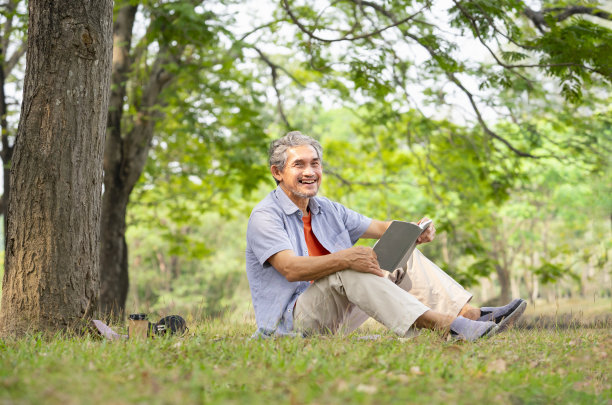 一位白发老人拿着一本书，面带微笑，坐在森林公园的一棵大树旁图片下载