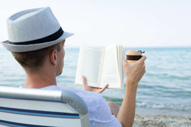 一个拿着咖啡杯的男人坐在沙滩上的躺椅上看书。图片下载