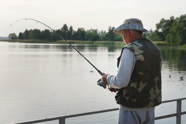 老人钓鱼。夕阳西下，一位白发苍苍的老渔夫从岸边抛下一缕丝线。积极的老年男性在湖中垂钓，旋转卷轴杆。退休渔民享受他的周末图片下载