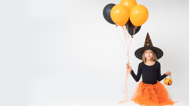 万圣节快乐的女孩戴着女巫帽，手里拿着一个南瓜形状的桶，桶里装着糖果和黑色和橙色的气球图片下载