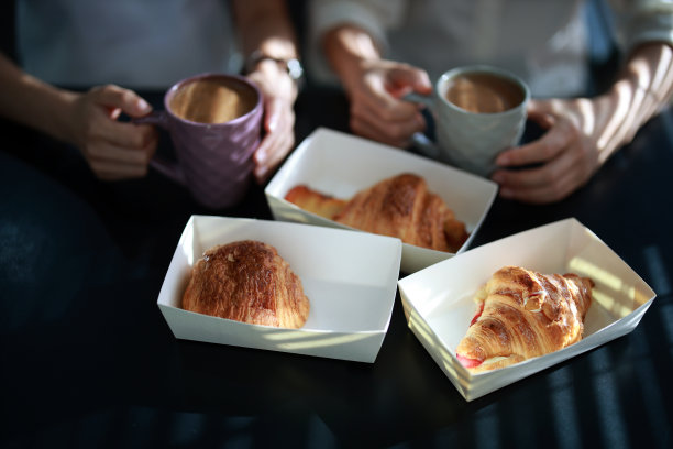 一张裁剪过的图片，两个年轻人在早晨享用着装在外卖纸盒里的咖啡和羊角面包，阳光照在桌子上。图片下载