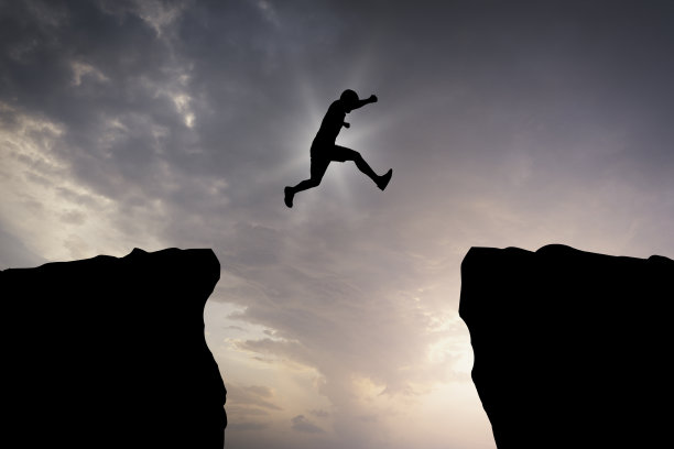 剪影的年轻人在日落时跳过两座落基山脉之间的悬崖。图片下载