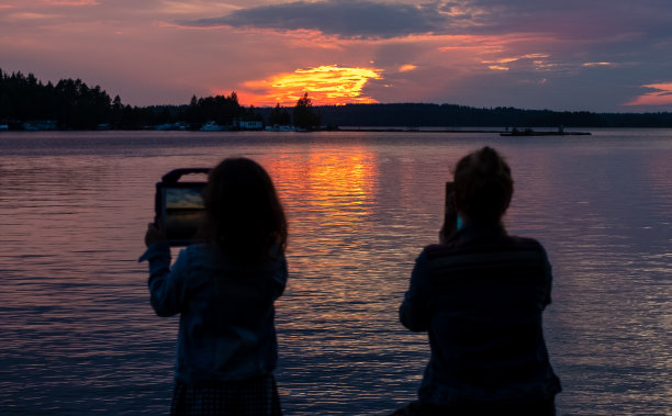 一个女孩，一个8岁的孩子，和一个年轻的女人在湖边看日落，用平板电脑拍照。图片下载