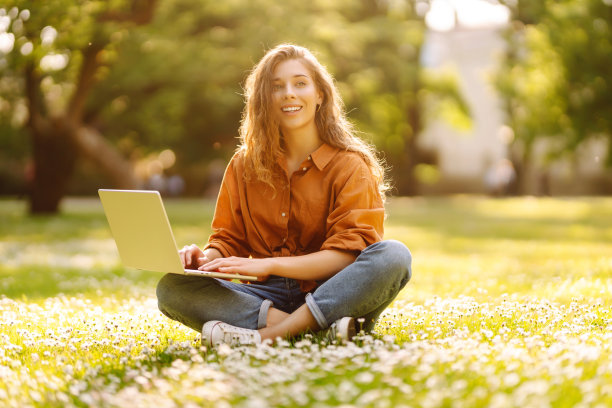一个年轻美丽的女性自由职业者坐在绿色的草地上，拿着一台笔记本电脑。图片下载