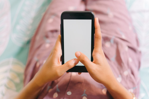 (特写)一个穿着睡衣的女人躺在床上，看着白色屏幕的智能手机。流媒体休闲概念图片下载