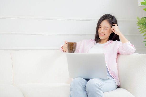 年轻的亚洲商业女性在家里用笔记本电脑工作，在客厅的沙发上喝咖啡，自由职业女孩坐在沙发上使用笔记本电脑，舒适放松，生活方式理念。图片下载