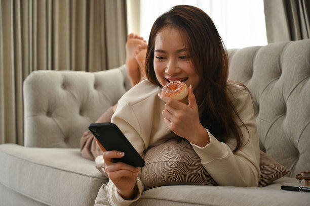 年轻的亚洲女子躺在沙发上玩手机吃甜甜圈的照片。图片下载