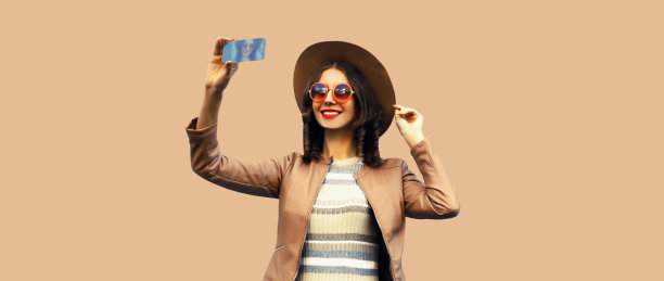 照片中的年轻时尚女子面带微笑，戴着圆帽，穿着棕色工作室背景的夹克，拿着手机自拍图片下载