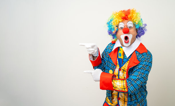 小丑先生。滑稽的喜剧演员的脸的肖像小丑在彩色制服指着文字广告的复制空间。快乐的表情让男呆子在孤立的背景下摆出各种姿势。图片下载