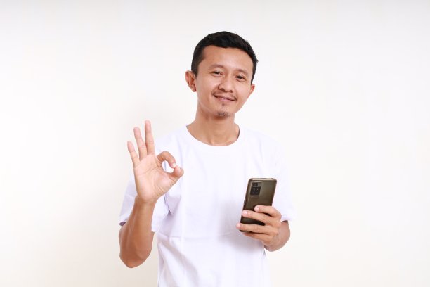 快乐的亚洲有趣的人拿着手机，同时显示好的手势。孤立于白色图片下载