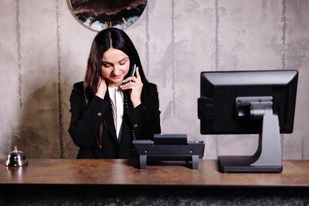 宾馆接待员。现代化的酒店前台，带铃。快乐的女接待员站在酒店柜台和电话交谈。图片下载