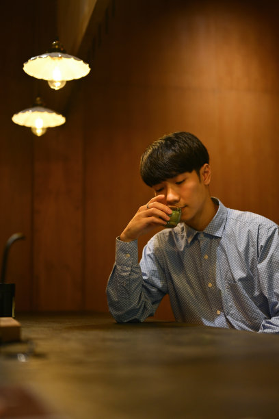 在温暖的光线下，年轻的亚洲男子在餐厅柜台的小茶杯里喝着热茶。图片下载