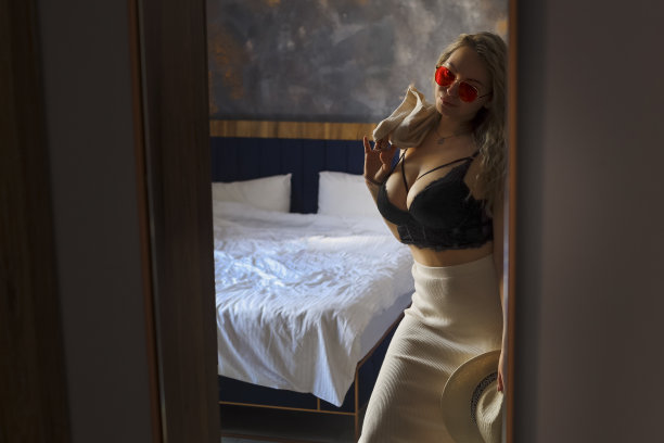 时髦漂亮的年轻女子，穿着优雅的衣服，戴着帽子，穿着胸罩和裙子，站在房间里的镜子旁边。美丽的金发女郎背对着镜头站着图片下载