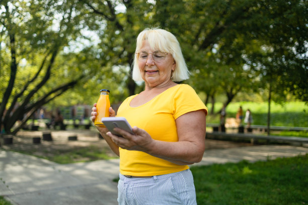 开朗的白种成熟女人妻子在公园里拿着一瓶果汁，拿着智能手机。高质量照片图片下载