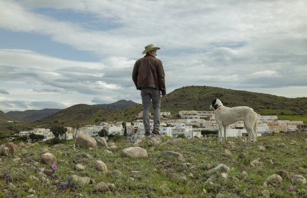 戴着牛仔帽的成年男子和他的狗站在田野上，城市在天空的背景下图片下载