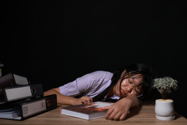 穿着校服，戴着眼镜，疲惫的年轻女学生坐在房间里一张不舒服的办公桌前睡觉。疲惫的女学生在晚上读了一长本书后休息。图片下载