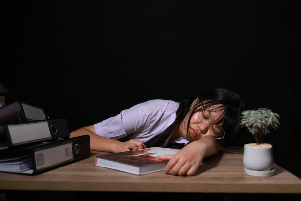 穿着校服，戴着眼镜，疲惫的年轻女学生坐在房间里一张不舒服的办公桌前睡觉。疲惫的女学生在晚上读了一长本书后休息。图片下载