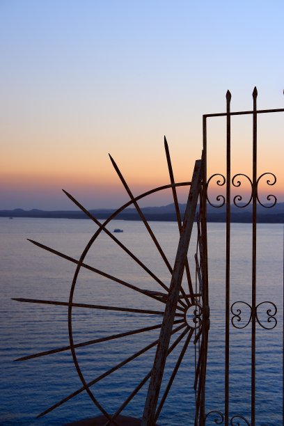 壮丽的山景和橙色的海上日落，通过锻造的镂空铁栅栏的花纹可以看到白色的船只图片下载