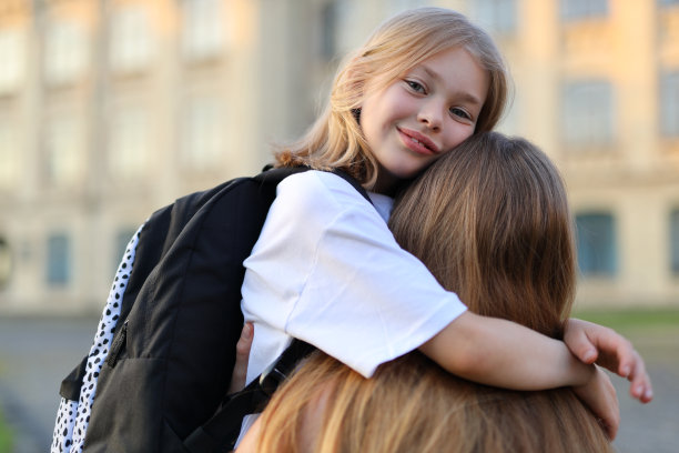 在一个阳光明媚的日子里，母亲抱着背着书包的女儿去上学。图片下载