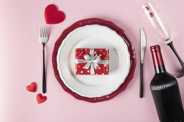 情人节浪漫的餐桌布置。礼物放在盘子里，粉红色背景的酒瓶和玻璃杯。餐厅约会，俯视图图片下载