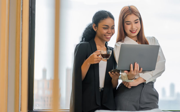 两个非洲和亚洲的混血美女，穿着休闲的衣服，开心的笑着，在室内办公室休息的时候聊天，看着笔记本电脑，端着咖啡。图片下载