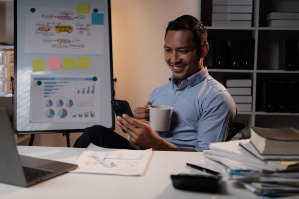 在办公室辛苦工作后，微笑的亚洲商人在办公桌前拿着智能手机和咖啡杯放松。图片下载