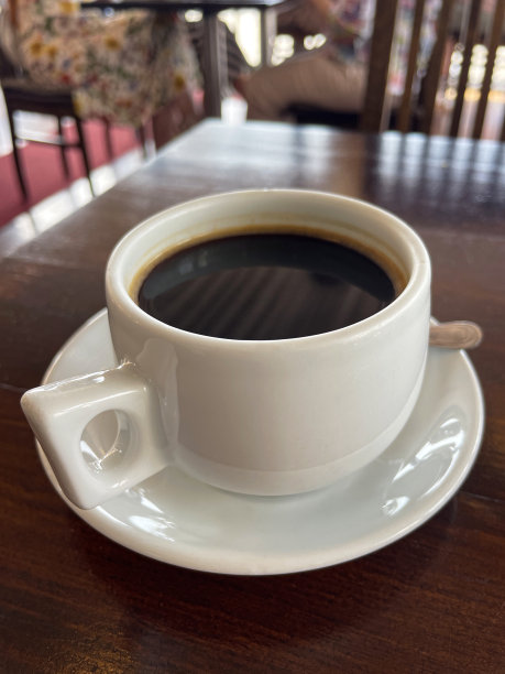 室内餐厅场景的特写图片，方形，木桌上摆放着白色茶杯和黑色美式咖啡碟，配有不锈钢茶匙，背景中有无法辨认的人物，重点放在前景上图片下载