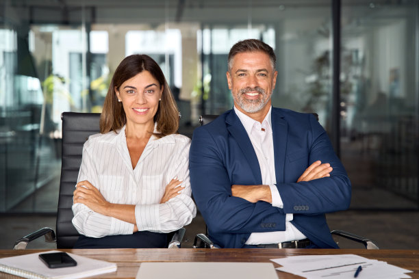 微笑的成熟的拉丁商人和欧洲商人坐在桌子旁，双臂交叉在办公室。两个不同的同事团队自信的专业商务人士。图片下载